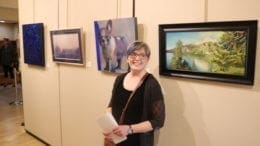 NancyJeanette Long smiling in front of her prizewinning painting, a portrait of a stocky dog with upright ears looking ahead skeptically