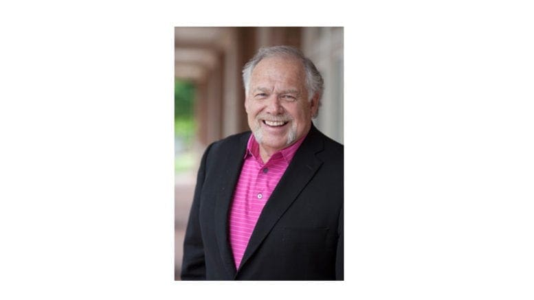 Max Bacon upper body shot. Bacon is bearded, with white hair. He is wearing a dark sports jacket and an open-collared dress shirt. The shot is an upper-body shot, and he is smiling face-forward into the camera