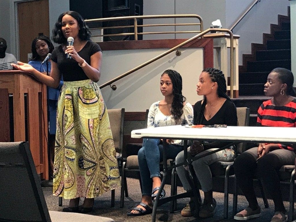 Student in black shirt and long yellow skirt speaking into a microphone.