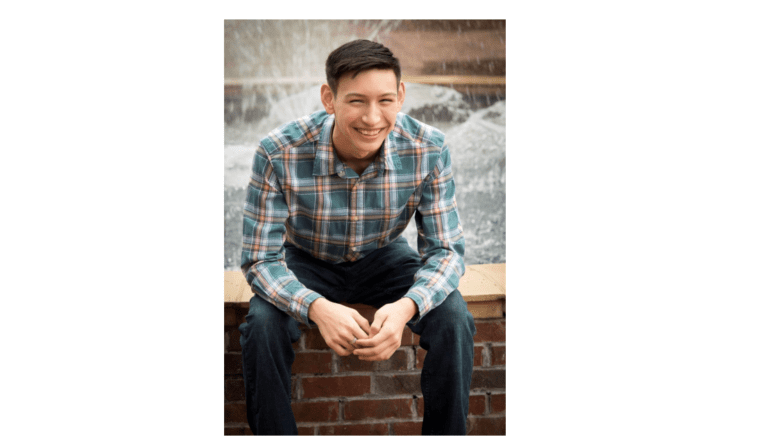 Austin Heller in plaid shirt seated on wall in front of fountain