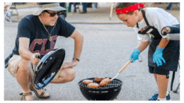 An adult male watching a child cook a piece of meat on a small grill