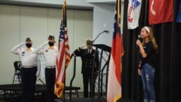 Three men in uniform salute flag as small girl sings