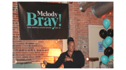 Photo of Melody Bray standing under one of her campaign banners, in front of a lamp and brick wall