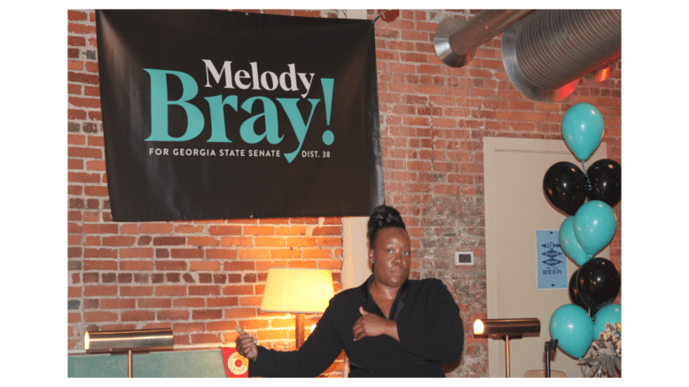 Photo of Melody Bray standing under one of her campaign banners, in front of a lamp and brick wall
