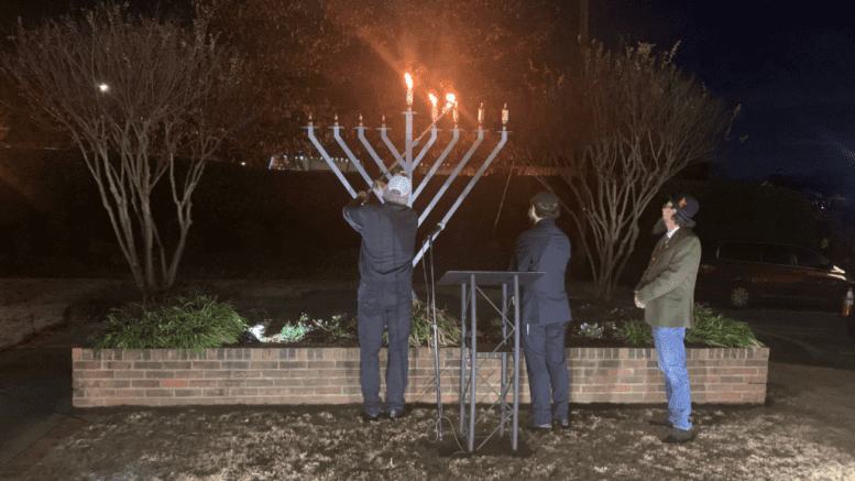 Three men standing in front of a Menorah . One of the men is lighting the third candle on the Menorah.