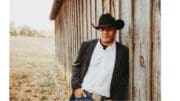 Marcus Flowers in black cowboy hat leaning against a vertical-paneled wooden barn wall