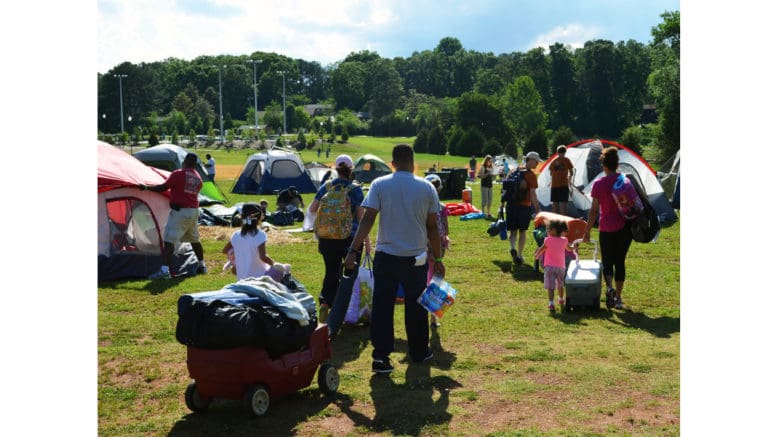 A group of families, backs to the camera, in the process of setting up a campsite with multiple tents