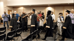 Students in caps and gowns enter room with family looking on