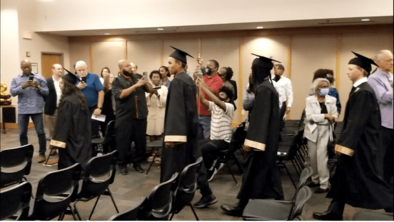 Students in caps and gowns enter room with family looking on