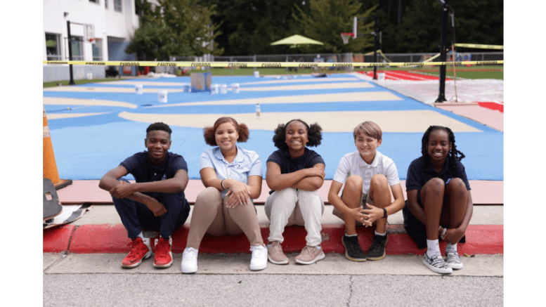 SAE students sit and smile in front of the coating being applied to the school’s basketball court and playground. Photo courtesy of Christopher Cavalier at TwoSenseFilms.