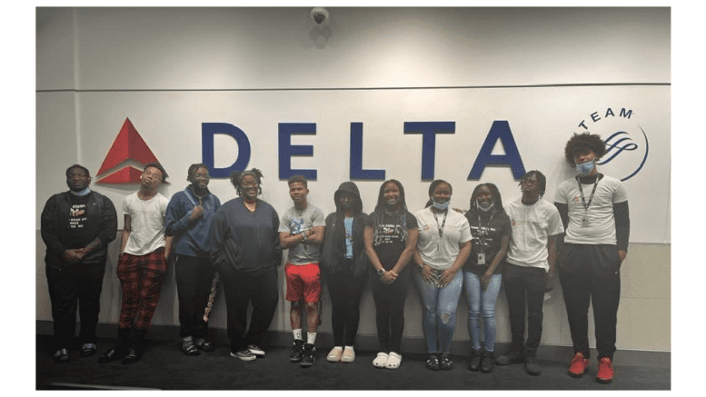 11 teens in front of a large DELTA Airlines sign