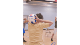 The older individuals pictured here, on the wooden floor of an indoor basketball court, were participating in an exercise class consisting of stretching, and aerobic repetitive motion movements. At this point in their exercise session, the participants were working the muscles of their arms and shoulders, by holding an inflatable volleyball with both hands, and lifting the ball over, and behind their head, resting it on their neck. This exercise also helped to stretch the muscles as well.