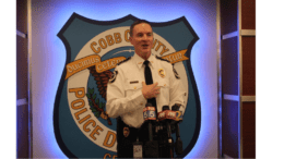 Cobb County Police Chief Stuart VanHoozer in front of large Cobb police badge emblem speaking toward camera with TV station microphones in front