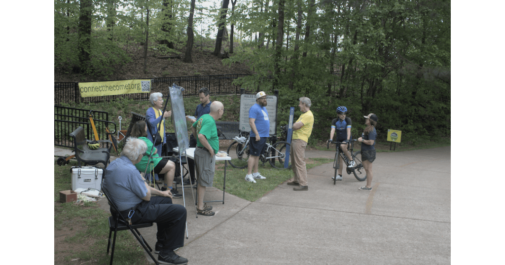 A group of people, one on a bicycle, stand around a table set up on the Silver Comet Trail