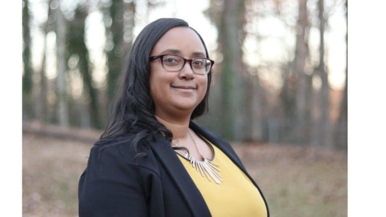 Laura Judge, wearing glasses, a light jacket and yellow blouse stands in front of a lightly wooded area