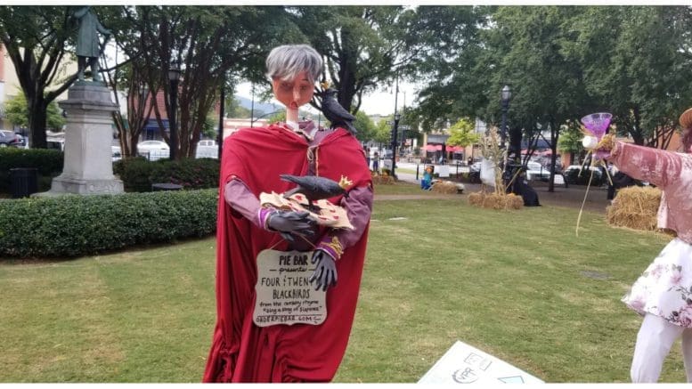 An elaborate scarecrow with a mannequin head holding a blackbird and a sign with a few words from the nursery rhyme (four and twenty blackbirds)