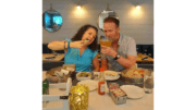 A man and woman in celebratory pose in restaurant, the woman moving a slice of pizza toward her mouth theatrically, the man holding a large drink