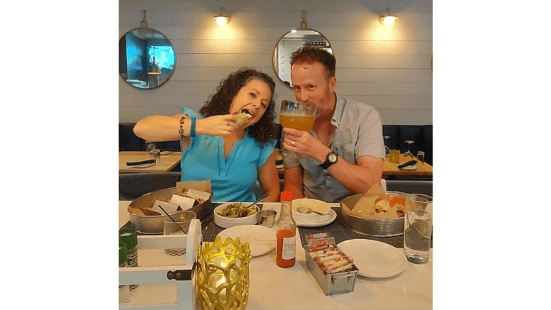 A man and woman in celebratory pose in restaurant, the woman moving a slice of pizza toward her mouth theatrically, the man holding a large drink