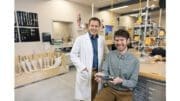 Mark Geil in a lab coat standing, and Harrison Bartlett leaning against a lab bench