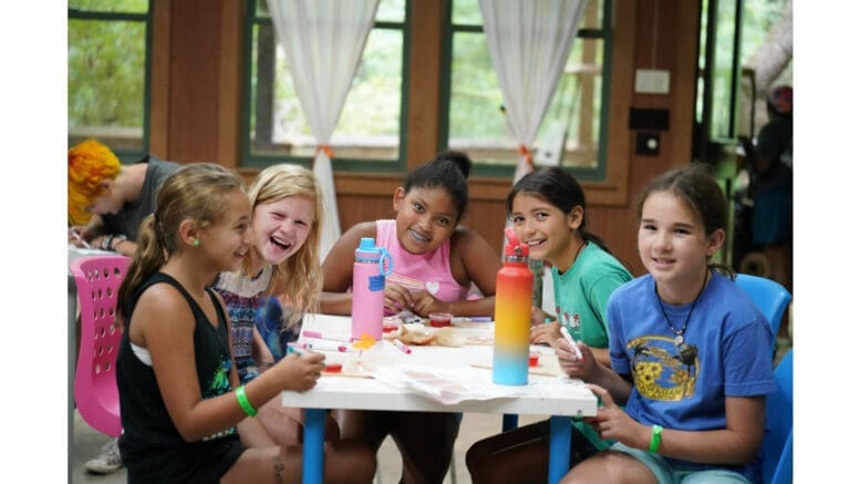 Several girls sit at a table with crafts in the Camp Timber Ridge dining hall