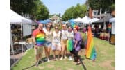 A group of men and women standing in a festival with tents, wearing rainbow-themed clothing