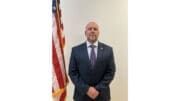 GBI Inspector Trent Hillsman wearing a dark suit and tie, poses with hands crossed in front next to an American flag