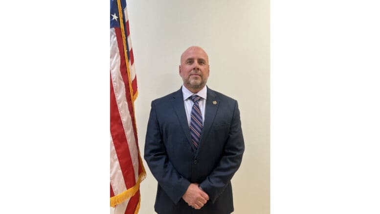 GBI Inspector Trent Hillsman wearing a dark suit and tie, poses with hands crossed in front next to an American flag