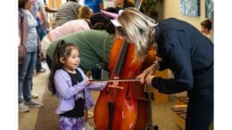 A little girl is near a string bass, and is being given a demonstration of how it works by an adult