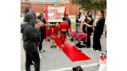 A red carpet leading to a sign that state "Light, Camera Action." A child in a superhero costume is on the carpet approaching the sign while bystanders applaud