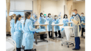 A group of nursing students in blue gowns in a semi-circle around a man who seems to be an instructor