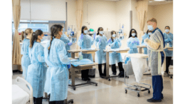 A group of nursing students in blue gowns in a semi-circle around a man who seems to be an instructor