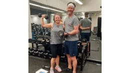 Woman and man standing by exercise equipment in a gym, smiling, woman raising dumbell in one hand