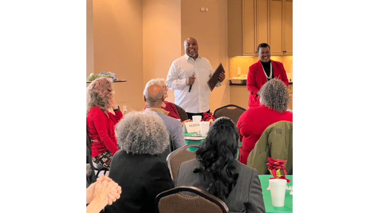 Man in a dress shirt with microphone in one hand and framed certificate speaks to assembled audience