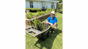 A smiling woman sits on the ground alongside a wheelbarrow of plants during a garden workday