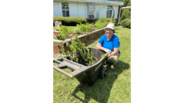 A smiling woman sits on the ground alongside a wheelbarrow of plants during a garden workday