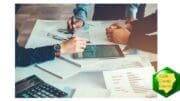 Photograph of three sets of hands working with a touchpad and calculator, documents and graphs on table before them