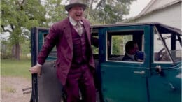A smiling man in a garish purple suit standing next to an early 20th Century auto