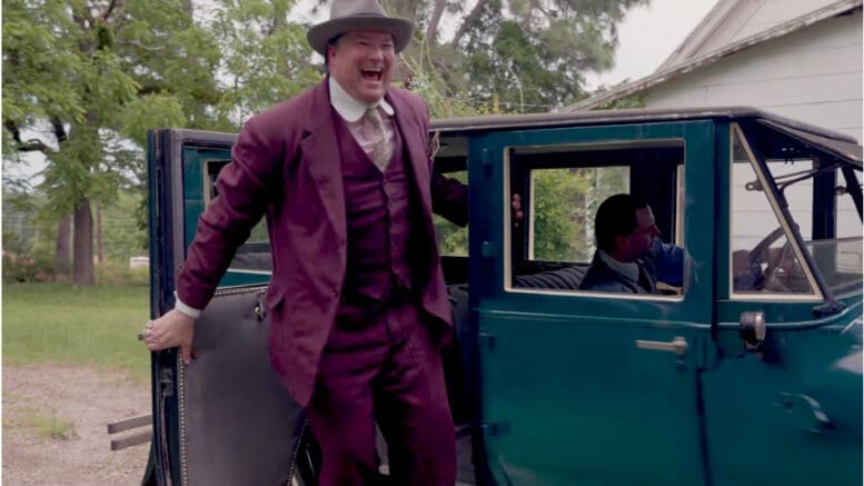 A smiling man in a garish purple suit standing next to an early 20th Century auto